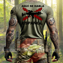 Cargar imagen en el visor de la galería, Hombre de espaldas con camiseta táctica color verde militar de Depredador Hispano en un entorno desértico. En el pecho lleva un logotipo con calavera táctica y casco, y en la espalda la frase “AQUÍ SE HABLA ESPAÑOL” junto a un fusil sobre fondo de Cruz de Borgoña. Ropa militar de alto rendimiento con diseño hispano.

