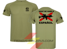 Cargar imagen en el visor de la galería, Camiseta táctica color verde militar de Depredador Hispano en un entorno desértico. En el pecho lleva un logotipo con calavera táctica y casco, y en la espalda la frase “AQUÍ SE HABLA ESPAÑOL” junto a un fusil sobre fondo de Cruz de Borgoña. Ropa militar de alto rendimiento con diseño hispano.
